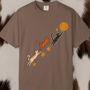 Peut inclure: Un t-shirt marron à manches courtes orné d'un motif représentant trois chats sautant en diagonale vers une lune orange ronde. Les chats sont de couleur crème, orange et noire, avec de petites étoiles orange à quatre branches.