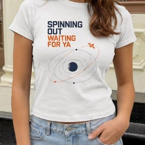Puede incluir: Una camiseta blanca de manga corta con el texto "SPINNING OUT WAITING FOR YA" en azul oscuro y naranja. Debajo del texto hay una ilustraci&oacute;n gr&aacute;fica de un planeta azul oscuro con finos anillos orbitales, peque&ntilde;os puntos y un sat&eacute;lite.