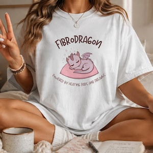 Könnte beinhalten: Ein weißes Kurzarm-T-Shirt mit einer Grafik eines kleinen rosa Drachen, der auf einem Kissen schläft. Der Text über dem Drachen lautet FIBRODRAGON und der Text darunter lautet POWERED BY HEATING PADS AND DREAMS in dunkelbrauner Schrift.