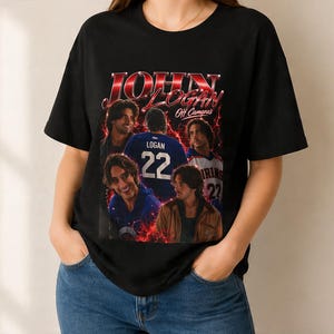 Pode incluir: Uma t-shirt preta de manga curta com uma impress&atilde;o gr&aacute;fica de John Logan e o texto 'JOHN LOGAN Off Camera'. O design inclui v&aacute;rias imagens de uma pessoa, uma camisola com o n&uacute;mero 22 e efeitos de rel&acirc;mpagos vermelhos na frente.