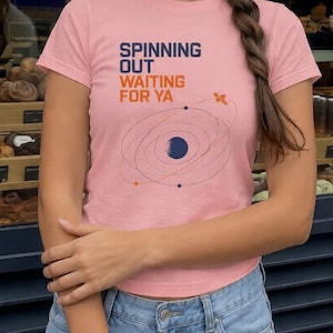 Puede incluir: Una camiseta de manga corta de color rosa claro con el texto "SPINNING OUT WAITING FOR YA" en azul oscuro y naranja. Debajo del texto hay una ilustraci&oacute;n gr&aacute;fica de un planeta azul oscuro con &oacute;rbitas, puntos de colores y un sat&eacute;lite.