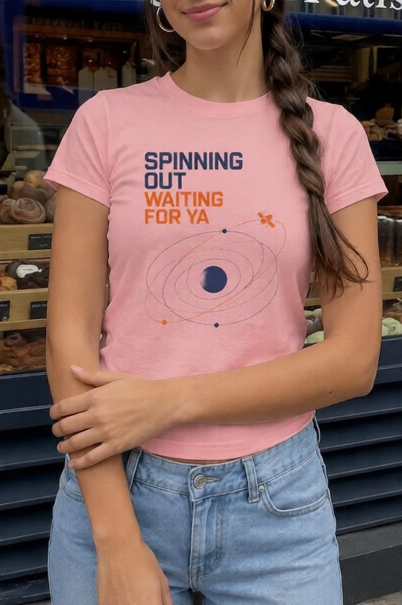 Puede incluir: Una camiseta de manga corta de color rosa claro con el texto "SPINNING OUT WAITING FOR YA" en azul oscuro y naranja. Debajo del texto hay una ilustraci&oacute;n gr&aacute;fica de un planeta azul oscuro con &oacute;rbitas, puntos de colores y un sat&eacute;lite.
