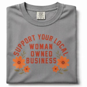 May include: A grey crew neck t-shirt featuring the orange text "SUPPORT YOUR LOCAL WOMAN OWNED BUSINESS" arched over the words in a block font. Two clusters of orange flowers with green stems and leaves frame the text on the left and right sides.
