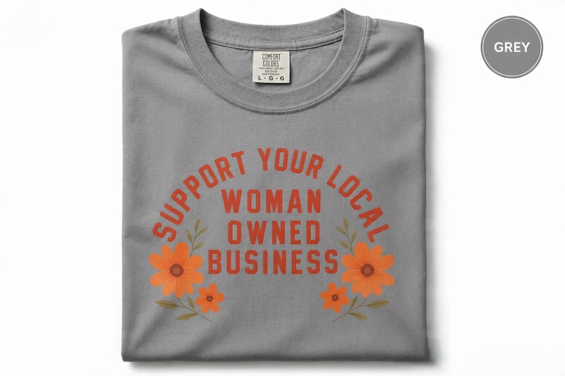 May include: A grey crew neck t-shirt featuring the orange text "SUPPORT YOUR LOCAL WOMAN OWNED BUSINESS" arched over the words in a block font. Two clusters of orange flowers with green stems and leaves frame the text on the left and right sides.