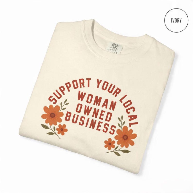 May include: An ivory crewneck t-shirt featuring the text SUPPORT YOUR LOCAL WOMAN OWNED BUSINESS in a rust-colored font. The design is arched and flanked by orange and rust-colored floral illustrations with green leaves on both sides.