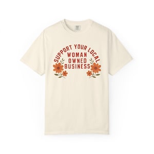 May include: A cream-colored short-sleeve t-shirt featuring the red text SUPPORT YOUR LOCAL WOMAN OWNED BUSINESS arched over the center. The design is flanked by two orange floral illustrations with green leaves on either side of the text.