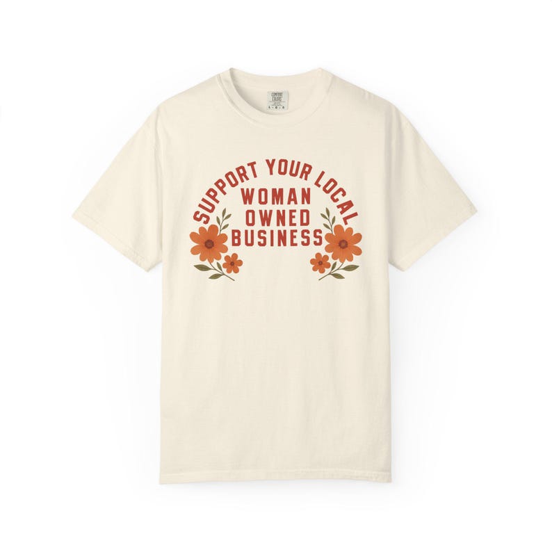 May include: A cream-colored short-sleeve t-shirt featuring the red text SUPPORT YOUR LOCAL WOMAN OWNED BUSINESS arched over the center. The design is flanked by two orange floral illustrations with green leaves on either side of the text.