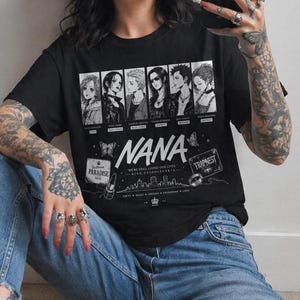 May include: A black short-sleeve t-shirt featuring a white graphic print of six manga characters from Nana. The design includes the text NANA, Trapnest, Paradise, and Tokyo, along with illustrations of butterflies, a lipstick, and a ring.