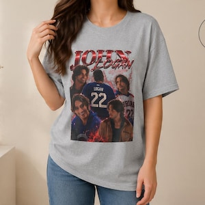 Pode incluir: Uma t-shirt cinzenta sport com uma impress&atilde;o gr&aacute;fica de John Logan e o texto "JOHN LOGAN" em letras vermelhas e pretas. O design inclui v&aacute;rias imagens da pessoa, incluindo uma com uma camisola azul com o n&uacute;mero 22.