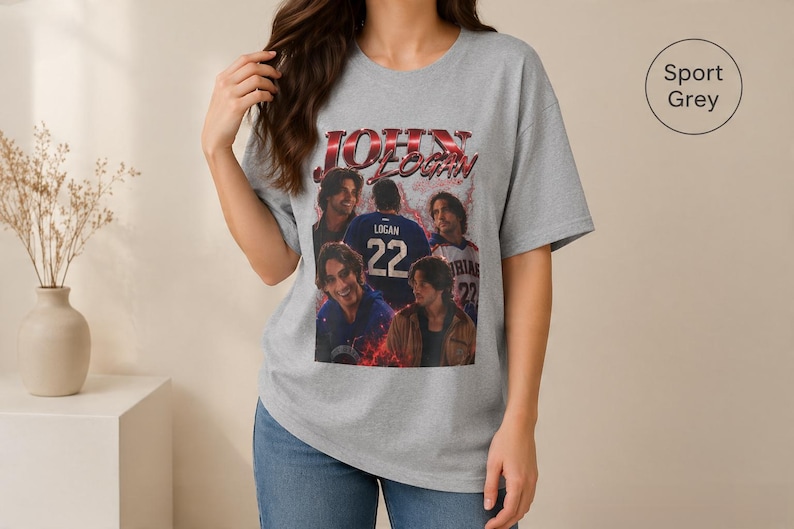 Pode incluir: Uma t-shirt cinzenta sport com uma impress&atilde;o gr&aacute;fica de John Logan e o texto "JOHN LOGAN" em letras vermelhas e pretas. O design inclui v&aacute;rias imagens da pessoa, incluindo uma com uma camisola azul com o n&uacute;mero 22.