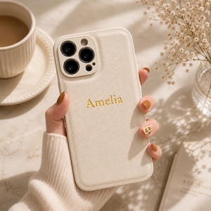 以下が含まれることがあります： クリーム色のテクスチャー加工が施されたスマートフォンケースで、金色の文字で「Amelia」という名前が刻印されています。ケースには3つの円形レンズとフラッシュ用の長方形のカメラカットアウトがあり、トリプルレンズカメラシステムを搭載したスマートフォンに対応しています。