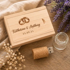 May include: A rectangular light-colored wooden box with a sliding lid featuring an engraved illustration of two interlocking rings, the names Katharine & Anthony, and the date 25.09.21. Beside it lies a cork-covered USB flash drive and a small glass vial.