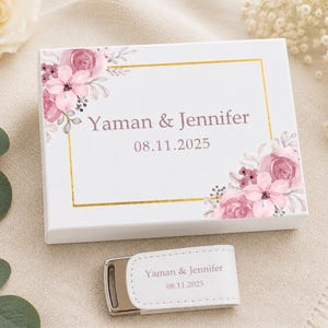 Può includere: Una scatola regalo rettangolare bianca e una chiavetta USB bianca abbinata. Entrambi gli articoli presentano una cornice dorata, illustrazioni floreali rosa e il testo "Yaman & Jennifer" seguito dalla data "08.11.2025" in un carattere rosa scuro.