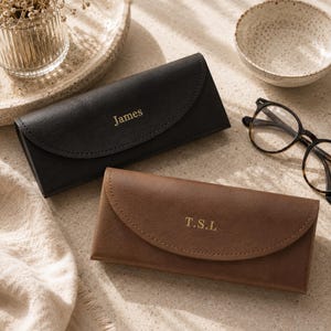 May include: A black rectangular glasses case with the name James embossed in gold and a brown rectangular glasses case with the initials T.S.L embossed in gold, both featuring a curved flap closure and a smooth, matte finish.