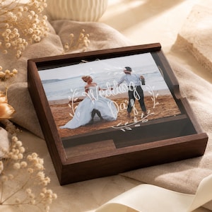 May include: A square dark wood display box with a clear acrylic lid featuring a printed photograph of a couple on a beach. The white text on the lid reads Wedding Day, framed by a delicate botanical wreath design, showcasing a keepsake frame.