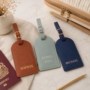 May include: Three rectangular luggage tags in rust, sage green, and navy blue, each featuring a matching strap with a metal buckle. The tags are personalised with the names SOPHIE, JAMES NYC, and MICHAEL embossed in a clean, sans-serif font.
