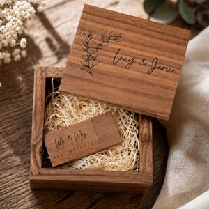 Op de afbeelding: Een vierkante houten doos met een afneembaar deksel voorzien van een lasergegraveerd botanisch ontwerp en de tekst Lucy & Jamie. Binnenin ligt een houten USB-stick in snipperpapier, gegraveerd met Mr & Mrs en de datum est. 03/07/21.