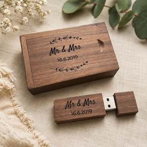 May include: A rectangular wooden box with a sliding lid and a matching wooden USB flash drive. Both items feature dark brown wood grain and are engraved with the text "Mr & Mrs" followed by the date "16.6.2019" and decorative leaf accents.