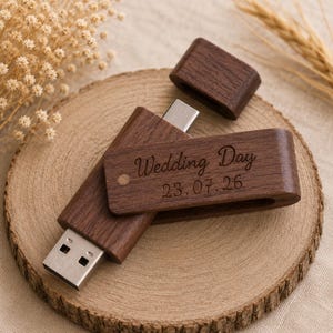 Może przedstawiać: Ciemnobrązowy drewniany pendrive z zdejmowaną nasadką. Na obudowie wygrawerowano napis Wedding Day oraz datę 23.07.26. Metalowe złącze USB jest widoczne na jednym końcu, a z boku znajduje się mały okrągły otwór.