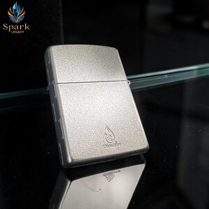 May include: A silver-colored metal lighter with a textured surface. The lighter has a rectangular shape with rounded edges and a hinged lid. The lighter features a flame logo and the text "Metal Fire".