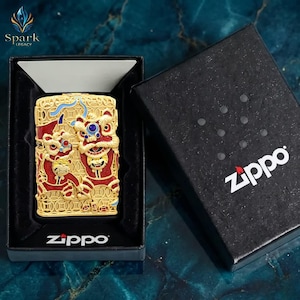 May include: Gold-colored Zippo lighter with a detailed relief of two Chinese lion dance figures in red, blue, and gold. The lighter is in a black box with the Zippo logo. The Spark Legacy logo is in the upper left corner.
