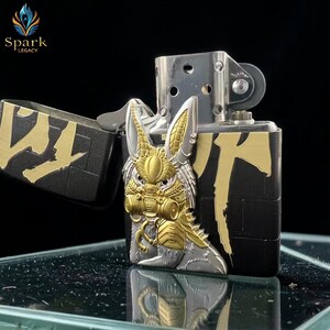 May include: A silver and gold-toned metal lighter with a detailed rabbit design on the front. The lighter has a black base with gold accents and the text "Spark Legacy" in the upper left corner.