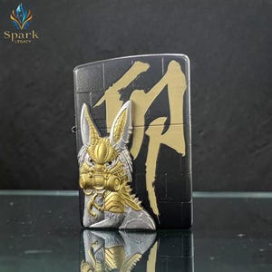 May include: Black and gold metal lighter with a detailed rabbit design on the front. The rabbit has a gas mask and armor. Gold Japanese characters are on the right side of the lighter. The brand "Spark Legacy" is in the upper left corner.
