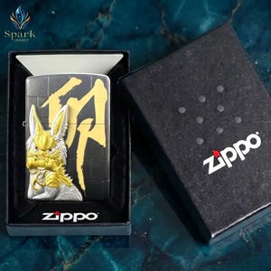 May include: A black Zippo lighter with a gold and silver design featuring a stylized creature and Japanese characters. The lighter is in a black box with the Zippo logo. The Spark Legacy logo is in the upper left corner.