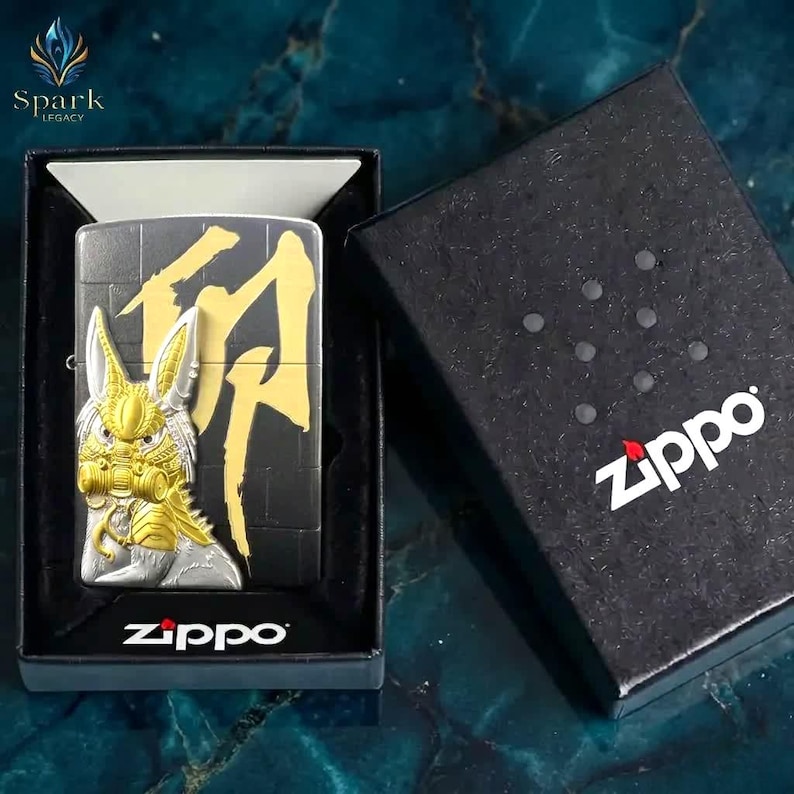 May include: A black Zippo lighter with a gold and silver design featuring a stylized creature and Japanese characters. The lighter is in a black box with the Zippo logo. The Spark Legacy logo is in the upper left corner.