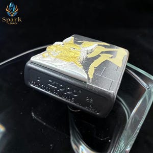 May include: A black Zippo lighter with a silver and gold design. The top of the lighter features a detailed relief of a figure with gold accents. The lighter's base is black, and the text "Zippo" is visible.