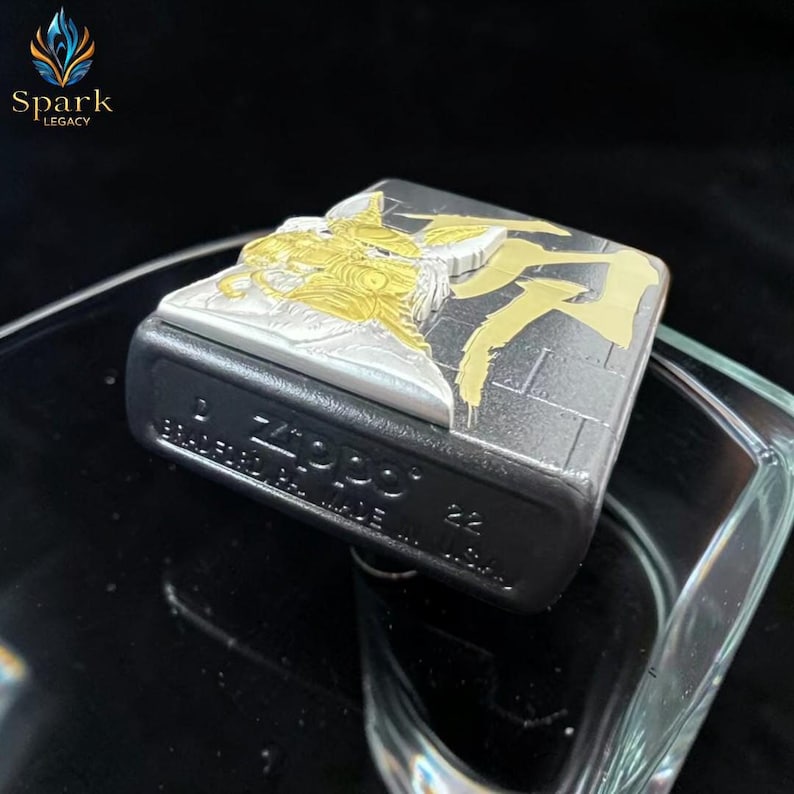 May include: A black Zippo lighter with a silver and gold design. The top of the lighter features a detailed relief of a figure with gold accents. The lighter's base is black, and the text "Zippo" is visible.