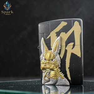 May include: A black and gold metal lighter with a detailed rabbit design on the front. The rabbit has gold accents and a gas mask. Gold Japanese characters are on the back. The brand "Spark Legacy" is in the upper left corner.