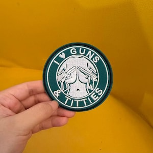 May include: A circular embroidered patch with a dark green background and a white border. The design features a white graphic of a torso with two handguns and the text I HEART GUNS & TITTIES in white capital letters around the center graphic.