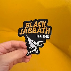 Könnte beinhalten: Ein schwarzer gestickter Aufnäher mit dem goldenen Schriftzug BLACK SABBATH und dem weißen Schriftzug THE END. Unter dem Text befindet sich eine weiße Stickerei einer geflügelten Figur in dynamischer Pose. Der Aufnäher hat einen schwarzen Rand.