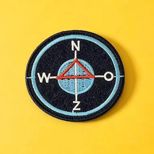 Puede incluir: Un parche bordado circular con fondo azul marino oscuro y un círculo interior azul claro. Un diseño de rosa de los vientos en blanco incluye las letras N, W y Z, con un triángulo rojo apuntando hacia arriba en el centro.