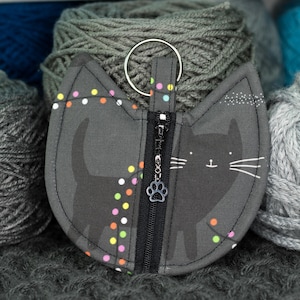May include: A gray cat-shaped zipper pouch with a paw print zipper pull. The pouch has a black cat silhouette and colorful confetti on a gray background.
