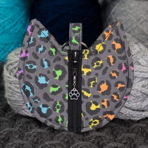 May include: A gray cat-shaped zippered pouch with a rainbow leopard print design. The pouch has a silver paw charm on the zipper pull.
