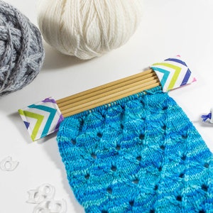 Puede incluir: Un proyecto de tejido a rayas azules y blancas en un juego de agujas de tejer de bambú con tapas de tela de colores. Las agujas descansan sobre una superficie blanca. Hay dos ovillos de lana, uno gris y otro blanco, y una pequeña bobina de hilo azul.