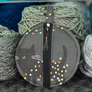 May include: A round, dark gray zippered pouch with a black cat design and a paw charm on the zipper. The pouch has a key ring and is surrounded by gray and blue yarn skeins. Colorful polka dots decorate the pouch.