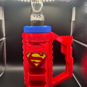 Puede incluir: Un "Assault Koozie" rojo y azul con el logo de Superman, sosteniendo una botella de vidrio. El koozie tiene un asa y una parte superior azul, con las palabras "Assault Koozie" impresas en el lateral. El logo de Superman es amarillo y rojo.