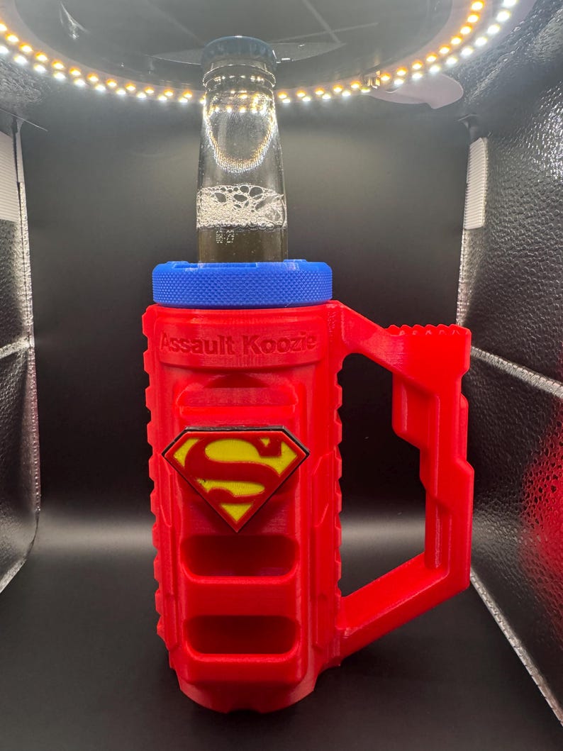 Puede incluir: Un "Assault Koozie" rojo y azul con el logo de Superman, sosteniendo una botella de vidrio. El koozie tiene un asa y una parte superior azul, con las palabras "Assault Koozie" impresas en el lateral. El logo de Superman es amarillo y rojo.