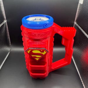 Puede incluir: Enfriador de latas rojo y azul con tem&aacute;tica de Superman, con asa. El enfriador tiene el logotipo de Superman y el texto "Assault Koozie". El borde superior es azul y el asa es roja. Dise&ntilde;ado para una lata est&aacute;ndar.