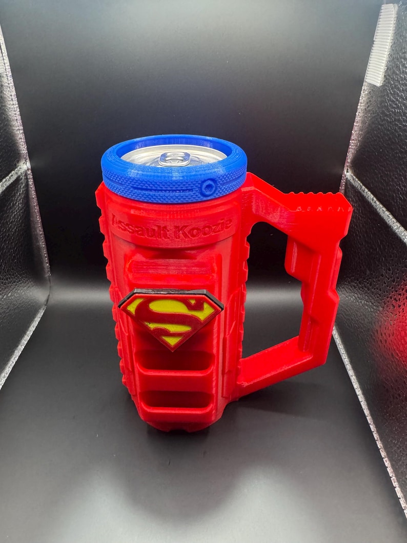 Puede incluir: Enfriador de latas rojo y azul con tem&aacute;tica de Superman, con asa. El enfriador tiene el logotipo de Superman y el texto "Assault Koozie". El borde superior es azul y el asa es roja. Dise&ntilde;ado para una lata est&aacute;ndar.