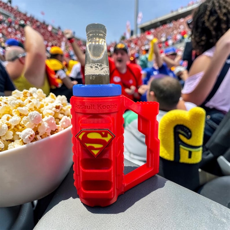 Puede incluir: Un Assault Koozie rojo con tem&aacute;tica de Superman con una tapa azul sostiene una botella de cerveza. El koozie tiene un logotipo de Superman amarillo y rojo. La imagen tambi&eacute;n muestra un cuenco de palomitas de ma&iacute;z y una multitud de personas en el fondo.