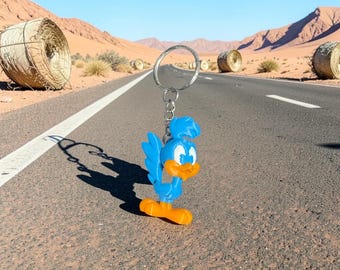 Puede incluir: Un llavero de plástico azul y naranja con forma de pájaro de dibujos animados con una cresta en la cabeza, unido a una anilla de metal plateado mediante una cadena corta. La figura presenta un cuerpo azul brillante, una mancha blanca y pico.