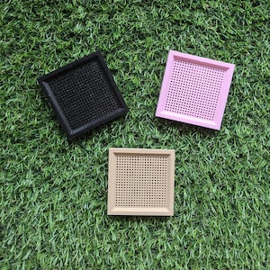May include: Three square trays with raised edges and a perforated grid pattern on the base. One tray is black, one is light pink, and one is beige. Each tray features a flat, solid border surrounding the central mesh-like surface area.