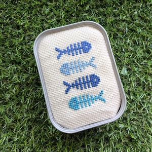Peut inclure: Une boîte rectangulaire en métal avec un couvercle en tissu beige orné d'un motif au point de croix représentant quatre arêtes de poisson dans des tons alternés de bleu foncé, bleu clair et bleu sarcelle, alignées verticalement.