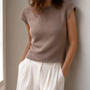 Può includere: Un top in maglia color tortora a maniche corte con scollo rotondo e un semplice motivo a maglia testurizzato. Il capo presenta un orlo dritto e maniche raglan, abbinato a pantaloni bianchi con un singolo bottone visibile in vita.
