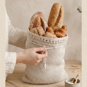 Può includere: Un sacco per il pane in maglia beige con coulisse e un bordo decorativo all'uncinetto sulla parte superiore. Il sacco è riempito con un assortimento di prodotti da forno dorati, tra cui una baguette lunga, pagnotte rotonde e croissant.