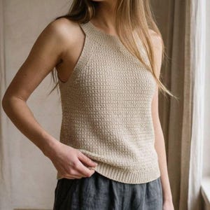 May include: A sleeveless beige knit tank top featuring a textured waffle stitch pattern, a high round neckline with ribbed trim, and a straight hemline. The garment is constructed with a consistent knit weave throughout the front and side panels.